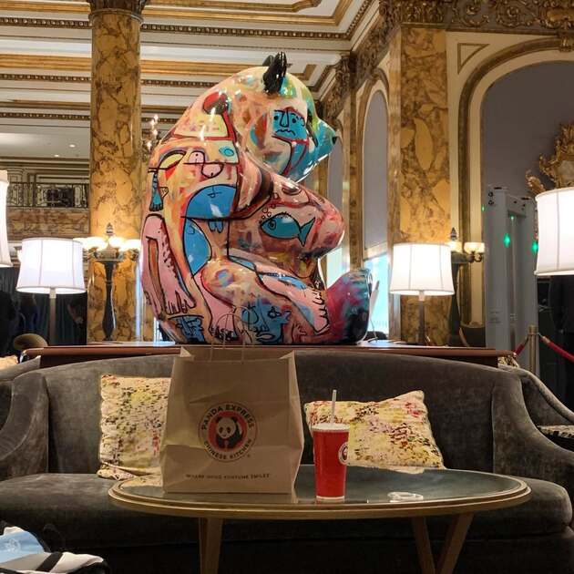 Hotel lobby with colorful panda sculpture on a large pedastal. Right is a metal detector. In front, a bag of Panda Express take out.